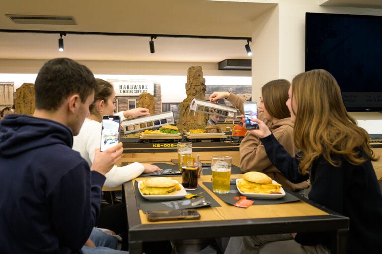 Customers seated at a table at Oliver Railway Restaurant receiving beers served by a miniature train, with retro American-style decorations on the wall.