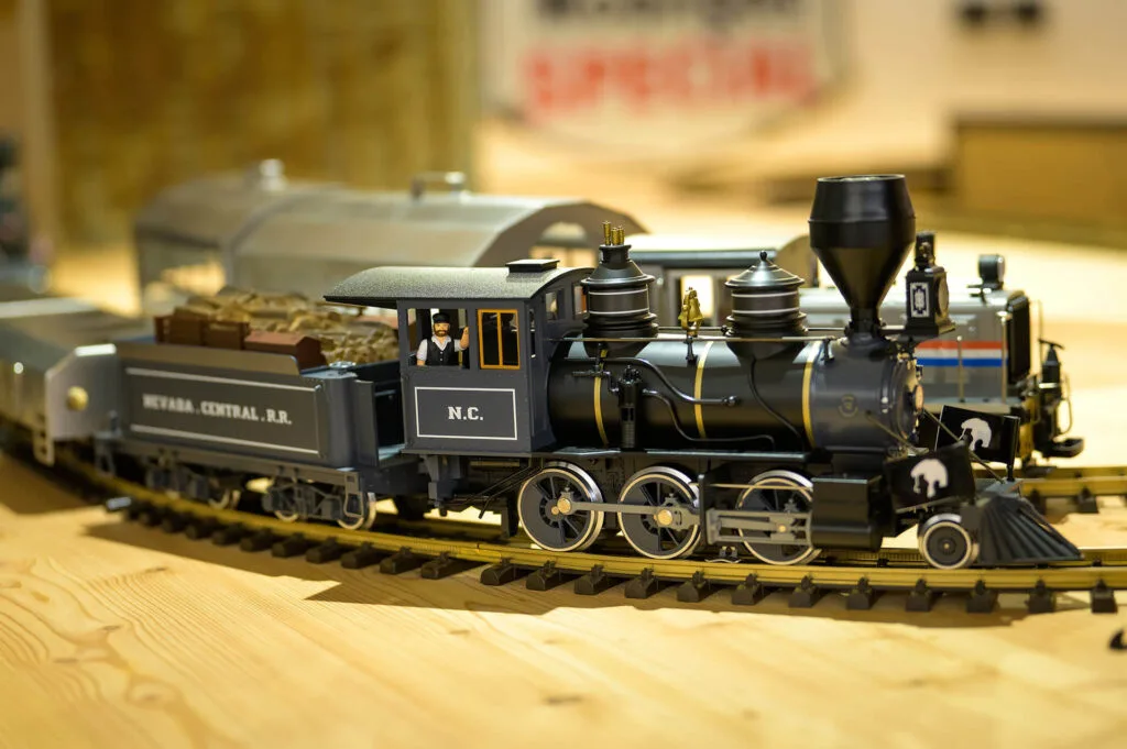 Black steam locomotive model with a coal wagon labeled Nevada Central R.R., featuring an American flag and miniature figures on board.