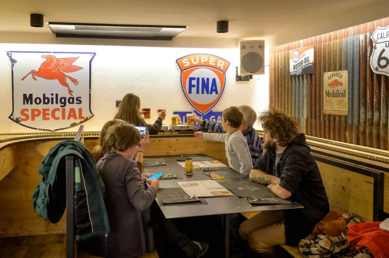 Customers seated at a table inside Oliver Railway Restaurant receiving beers delivered by a miniature train, with retro American-style decorations on the wall.