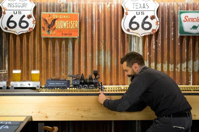 Uomo fotografa con lo smartphone un trenino elettrico che trasporta birre su binari davanti a parete in stile vintage